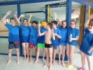 Neun Jugendliche mit blauen T-Shirts stehen im Schwimmbad. Sie jubeln. Ein Kind hält einen Pokal.